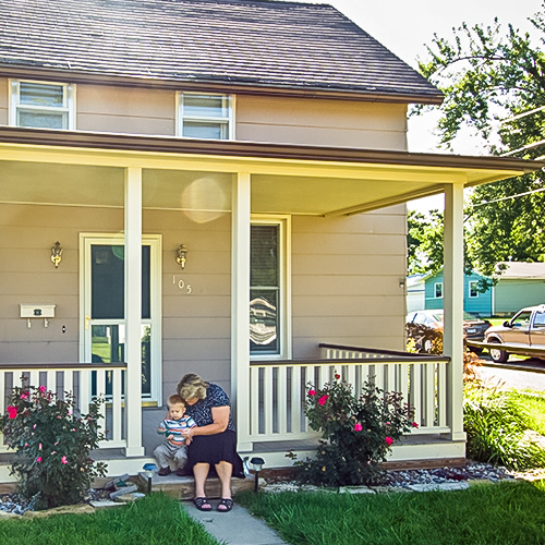 Woman holds a small child and sits on the front porch step of a quaint home